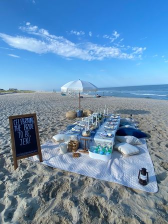 Beachside bridal picnic on a sunny sandy shore with a low table, cushions, umbrella, candles and place settings, chilled drinks in a tub and a chalkboard sign celebrating the bride-to-be, ocean waves and blue sky in the background
