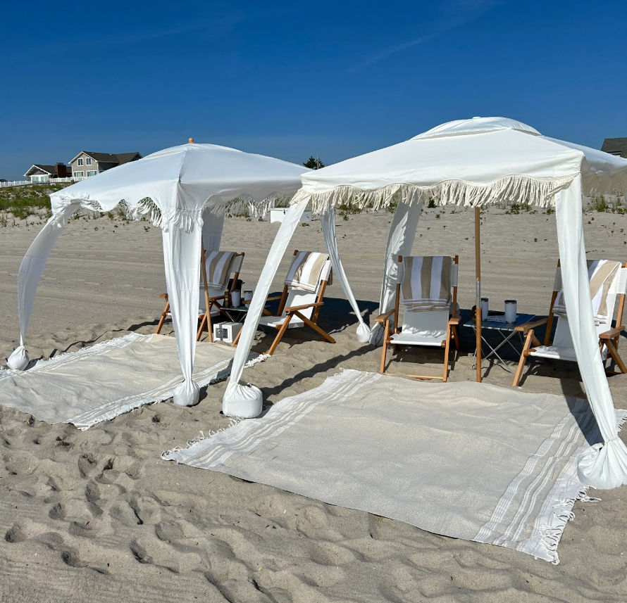 Sunny sandy beach with two white fringe canopy tents, striped wooden lounge chairs on woven rugs, a small side table, and coastal houses on the dunes under a clear blue sky.