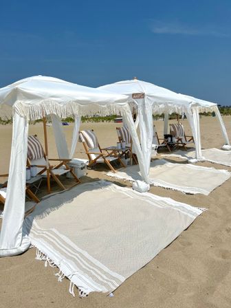 Sunny seaside scene with white fringe canopies and striped wooden lounge chairs set on sandy shore under a clear blue sky