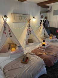 Three white canvas teepees for a kids' indoor slumber party in a cozy living room, with blush blankets, pillows, small trays holding lanterns and snacks, warm string lights and rustic wall decor.