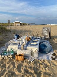 Coastal picnic on the New Jersey shore — low white table set on sand with blue-gray linens, candles, plates, sparkling water bottles, cushions and a small chalkboard sign, with dunes and ocean waves in the background.
