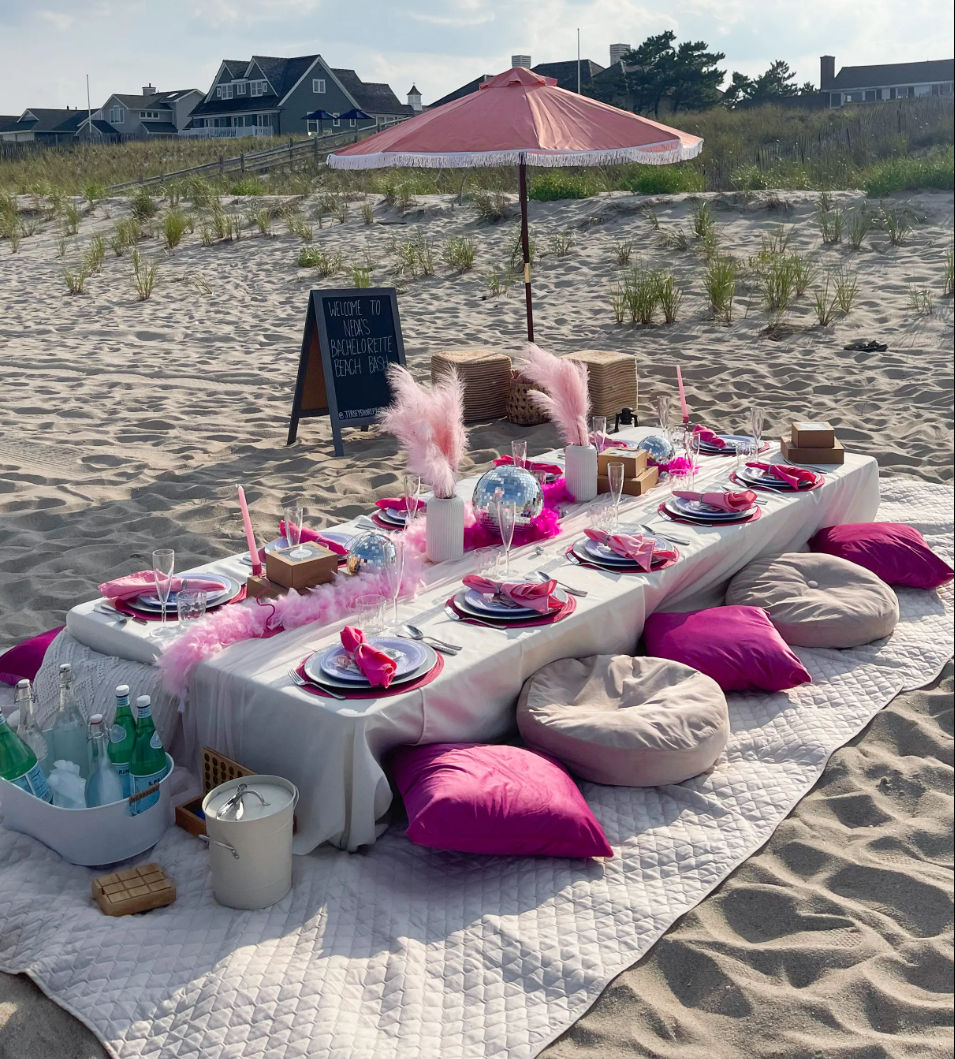 Coastal beach picnic on sandy dunes: low white table under a pink fringe umbrella, hot-pink pillows and round cushions on quilts, pink-feather centerpieces, disco balls, place settings and champagne flutes.