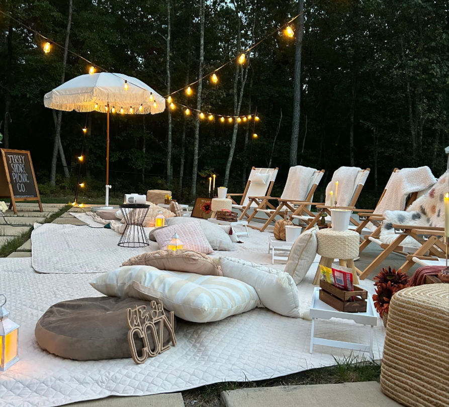 Evening backyard picnic lounge with quilted rugs, plush cushions and low wooden chairs under string lights and a fringed umbrella, lit by lanterns and candles against a wooded backdrop.