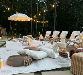 Evening backyard picnic lounge with quilted rugs, plush cushions and low wooden chairs under string lights and a fringed umbrella, lit by lanterns and candles against a wooded backdrop.