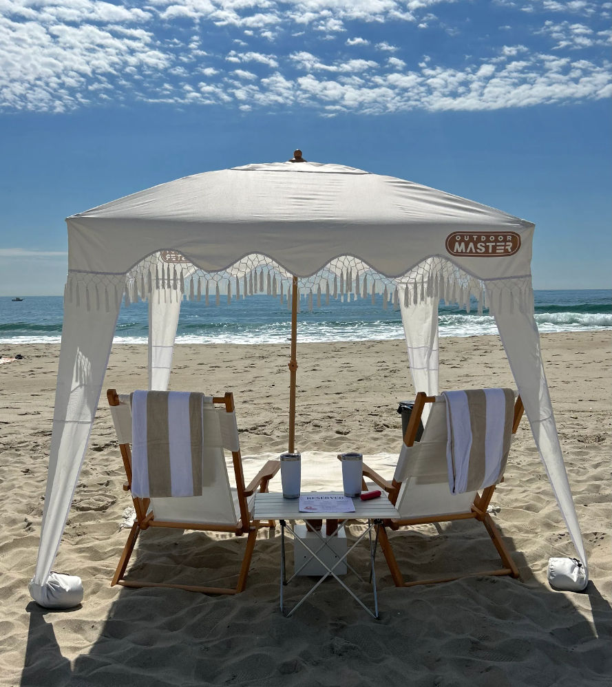 White fringed beach canopy shading two wooden lounge chairs with striped towels and a small table holding tumblers and a magazine on a sunny sandy oceanfront under a blue sky with scattered clouds.