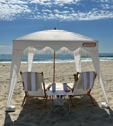 White fringed beach canopy shading two wooden lounge chairs with striped towels and a small table holding tumblers and a magazine on a sunny sandy oceanfront under a blue sky with scattered clouds.