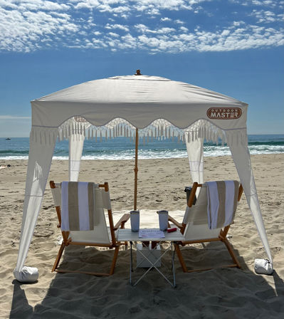 White fringed beach canopy shading two wooden lounge chairs with striped towels and a small table holding tumblers and a magazine on a sunny sandy oceanfront under a blue sky with scattered clouds.