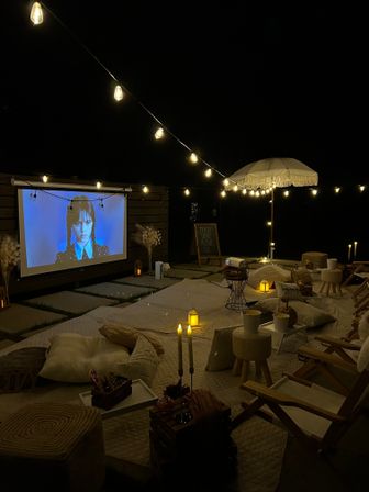 Cozy outdoor movie night on a backyard patio with a projected screen, warm string lights and lanterns, fringed umbrella, low picnic-style seating, cushions and candles creating a boho atmosphere