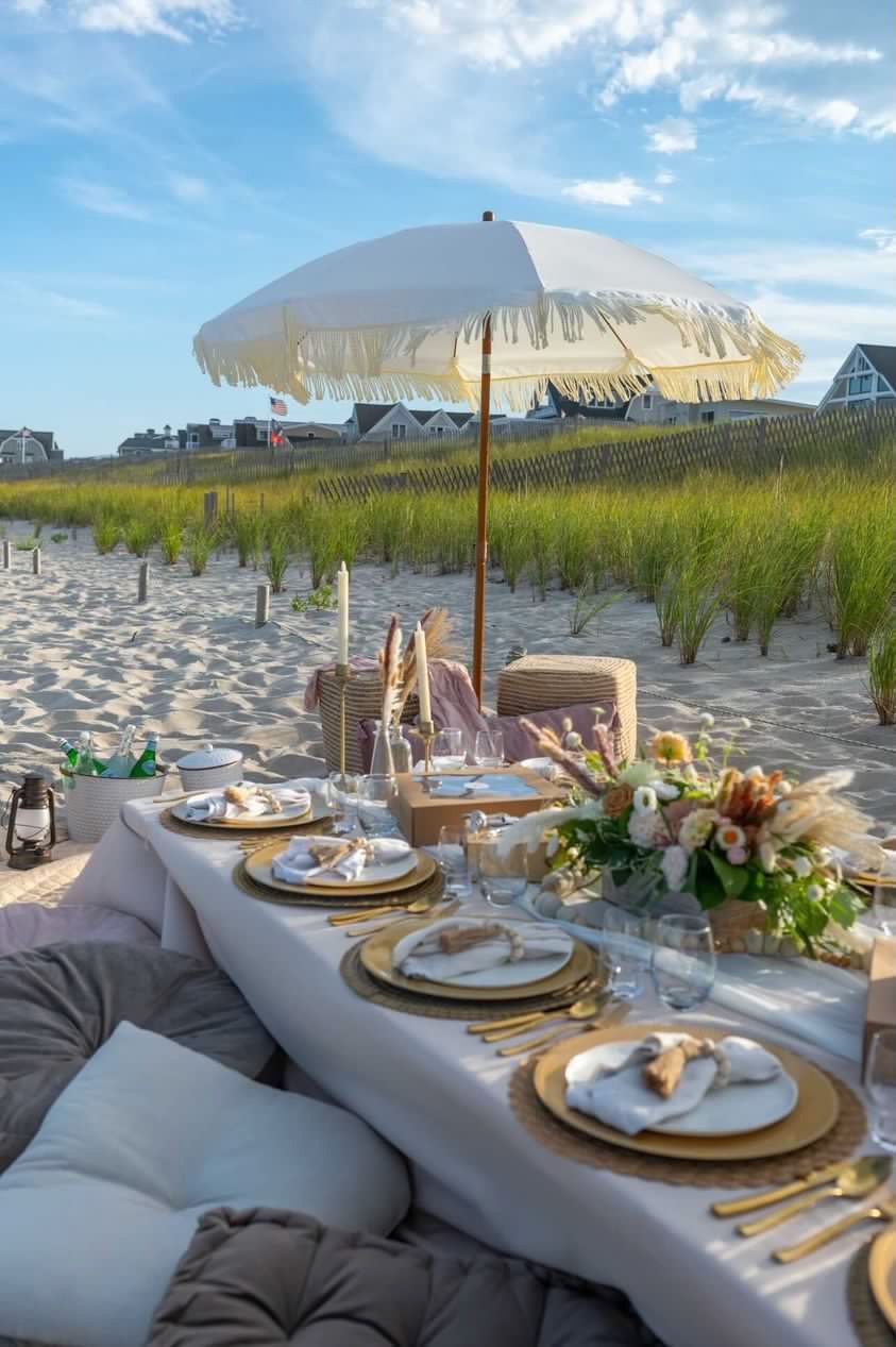 Boho beach picnic on a sandy coastal dune with a fringed umbrella, low table set with gold flatware, floral centerpiece and cushions