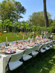 Al fresco backyard garden table set for a spring/summer party with white folding chairs, pink napkins, candles, glassware and hanging flowers over a green lawn.