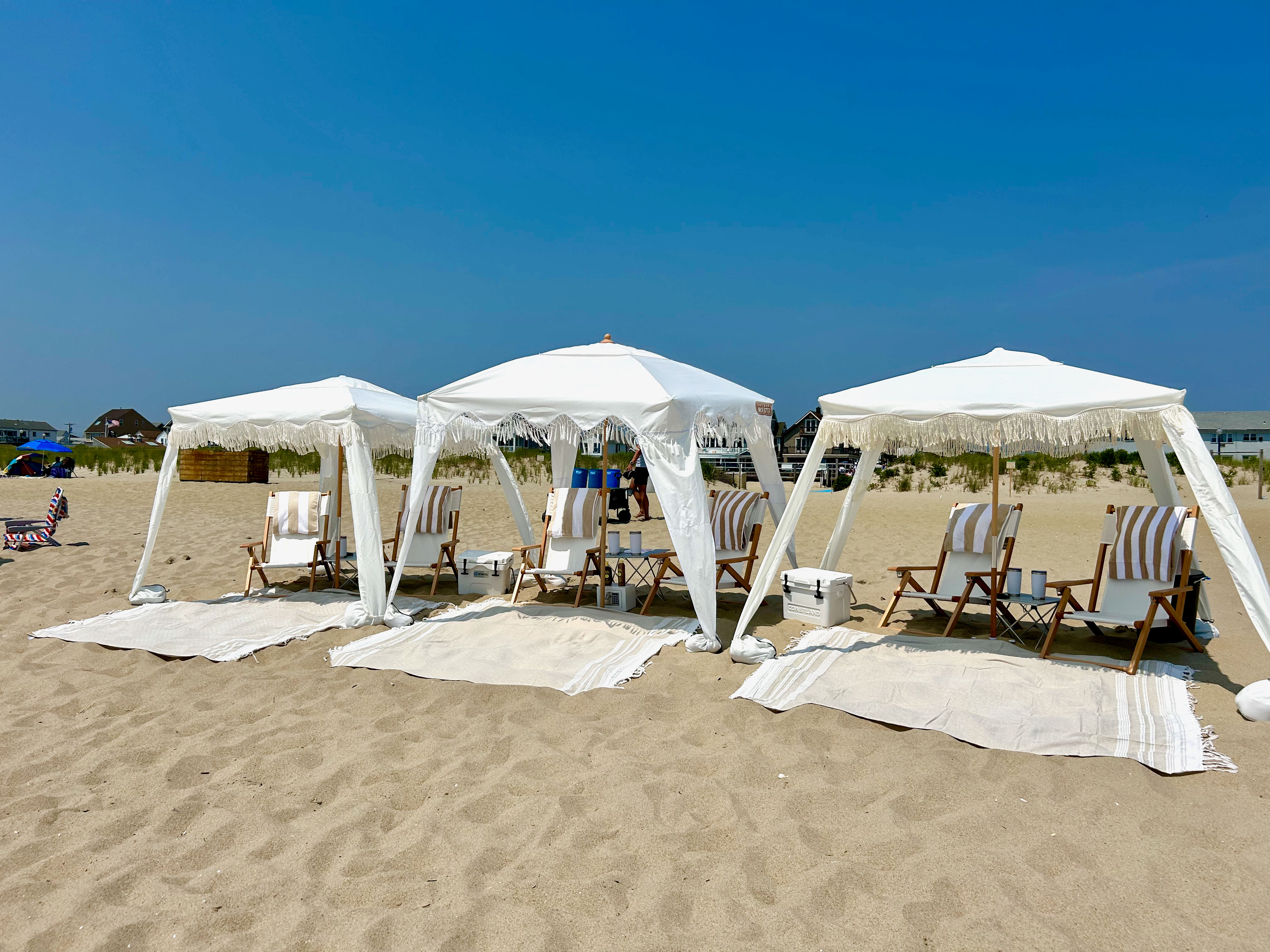 Three white fringe cabanas with wooden striped lounge chairs, coolers and rugs arranged on a sunny sandy beach under a clear blue sky.