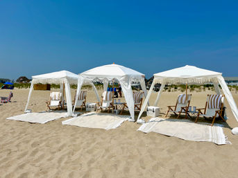 Three white fringe cabanas with wooden striped lounge chairs, coolers and rugs arranged on a sunny sandy beach under a clear blue sky.