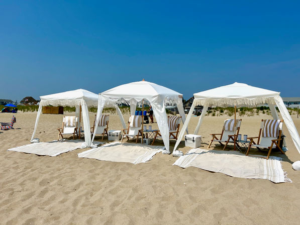 Three white fringe cabanas with wooden striped lounge chairs, coolers and rugs arranged on a sunny sandy beach under a clear blue sky.