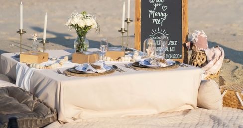 Romantic beach picnic proposal setup with a low linen table, candles, white roses, place settings and a chalkboard reading “Will you marry me?”