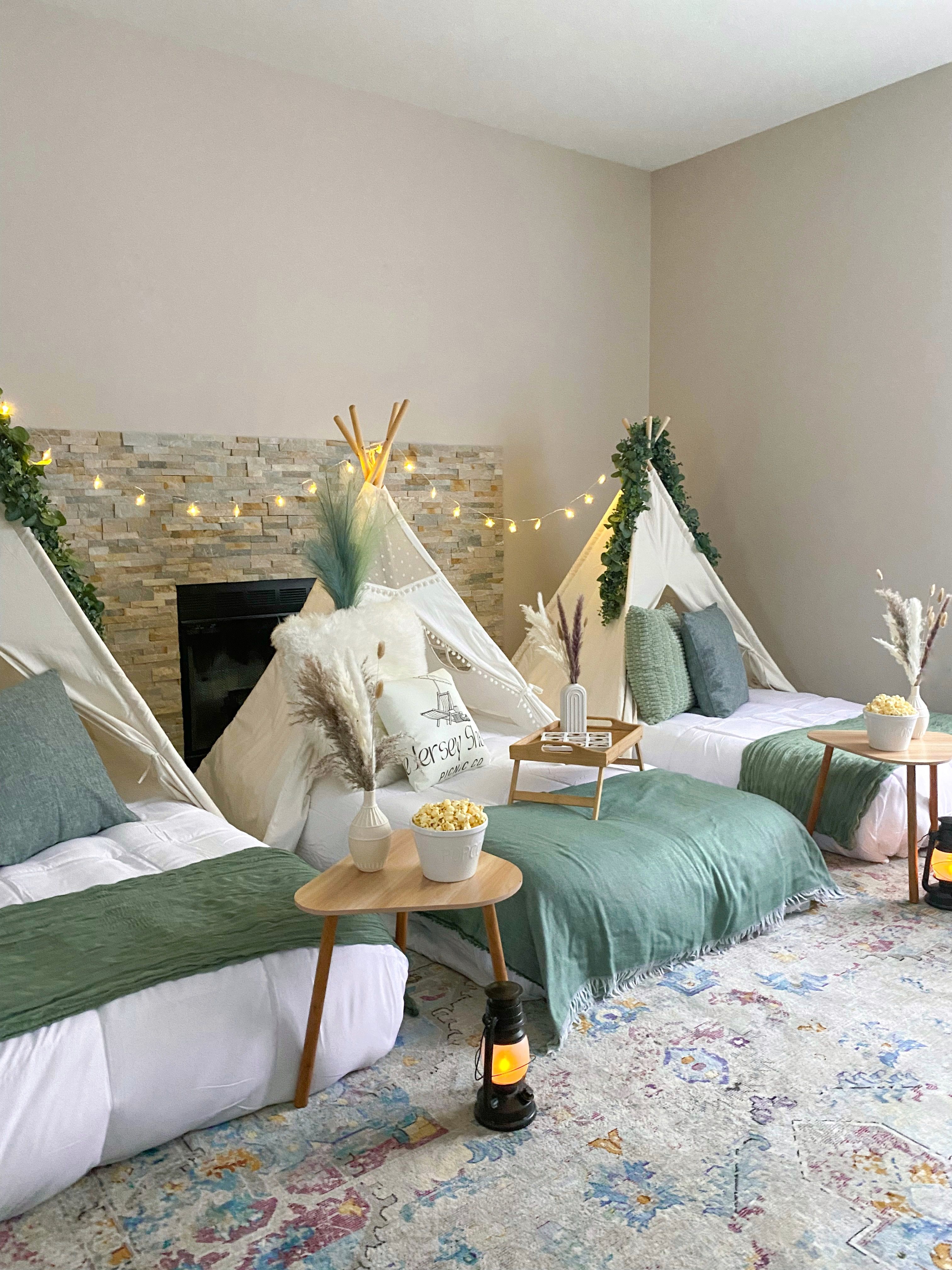 Cozy indoor slumber party in a living room with three white canvas teepees, low beds layered with green throws and pillows, wooden side tables holding popcorn bowls and vases of pampas grass, string lights and lanterns on a patterned rug.