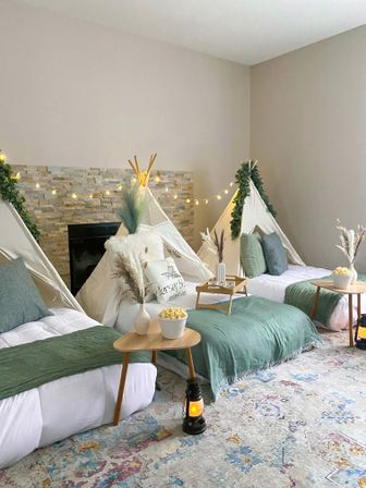 Cozy indoor slumber party in a living room with three white canvas teepees, low beds layered with green throws and pillows, wooden side tables holding popcorn bowls and vases of pampas grass, string lights and lanterns on a patterned rug.