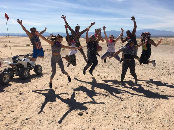 Energetic group jumping in mid-air on sandy desert dunes during an ATV off-road adventure, helmets and mountain skyline in the background.