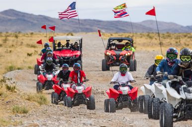 Group of ATV and UTV riders on a rocky desert trail, red and white quads and two off-road UTVs, riders wearing helmets and goggles, American and colorful flags flying overhead — energetic off-road adventure in an arid landscape