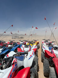 Row of colorful off-road ATVs parked in a sandy desert staging area, tall red safety flags fluttering under a clear blue sky with distant mountains and a trailer in the background.
