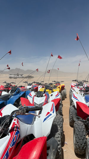 Row of colorful off-road ATVs parked in a sandy desert staging area, tall red safety flags fluttering under a clear blue sky with distant mountains and a trailer in the background.