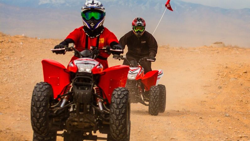 Helmeted riders on red ATVs kicking up dust on an off-road desert trail with distant mountains in the background
