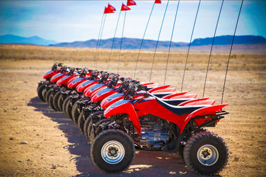 Bright red ATVs lined up on a sandy desert plain with tall red safety flags, ready for off‑road rides against distant mountains under a clear sky.