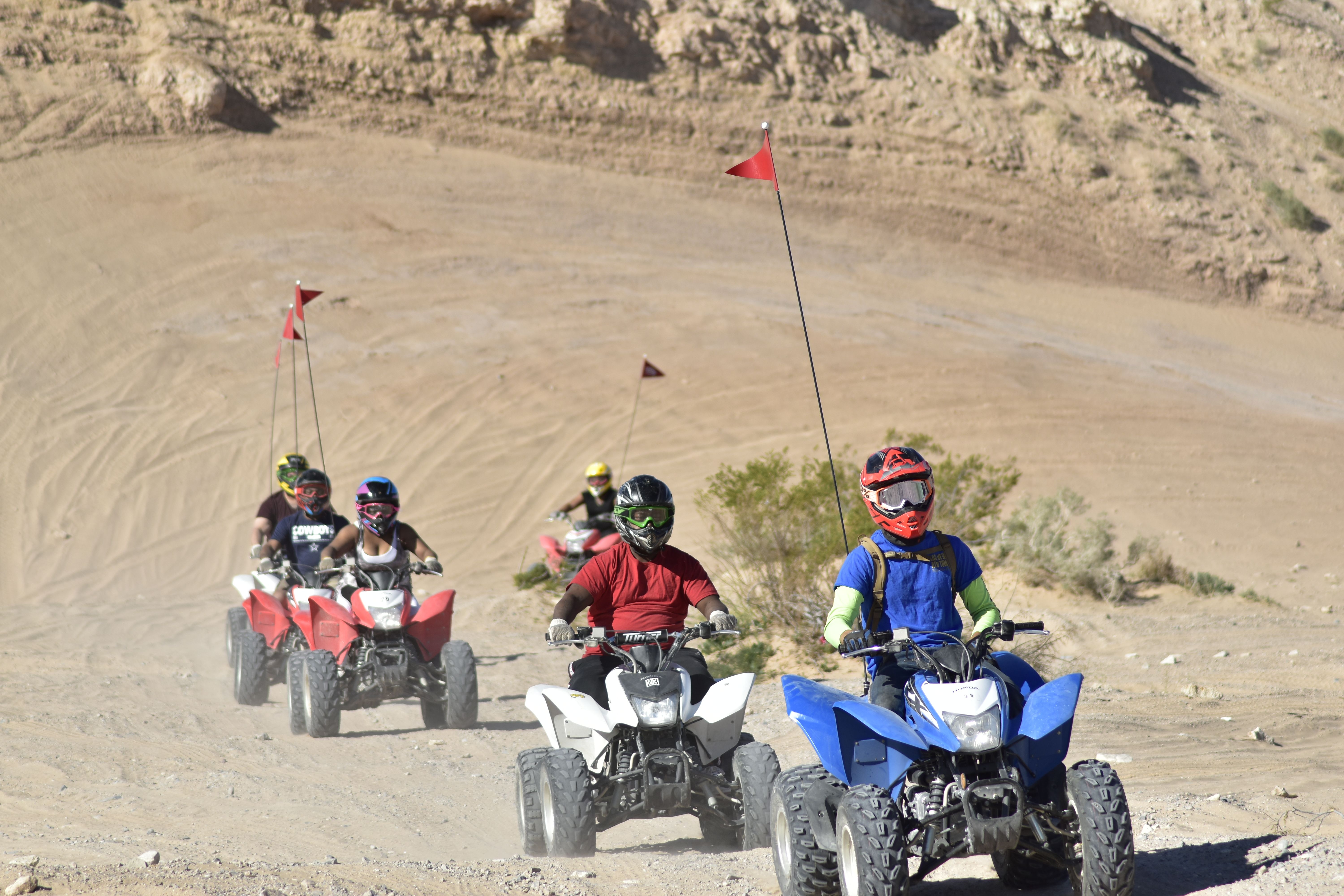 Group of ATV riders in colorful helmets on blue, white and red quad bikes with tall safety flags riding across a sandy desert dune trail