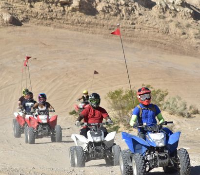 Group of ATV riders in colorful helmets on blue, white and red quad bikes with tall safety flags riding across a sandy desert dune trail