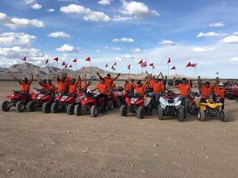 Cheerful group of ATV riders in orange shirts posing on red and white four-wheelers with tall red safety flags across a sandy desert with rocky mountains and a blue sky.