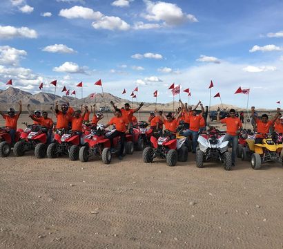 Cheerful group of ATV riders in orange shirts posing on red and white four-wheelers with tall red safety flags across a sandy desert with rocky mountains and a blue sky.