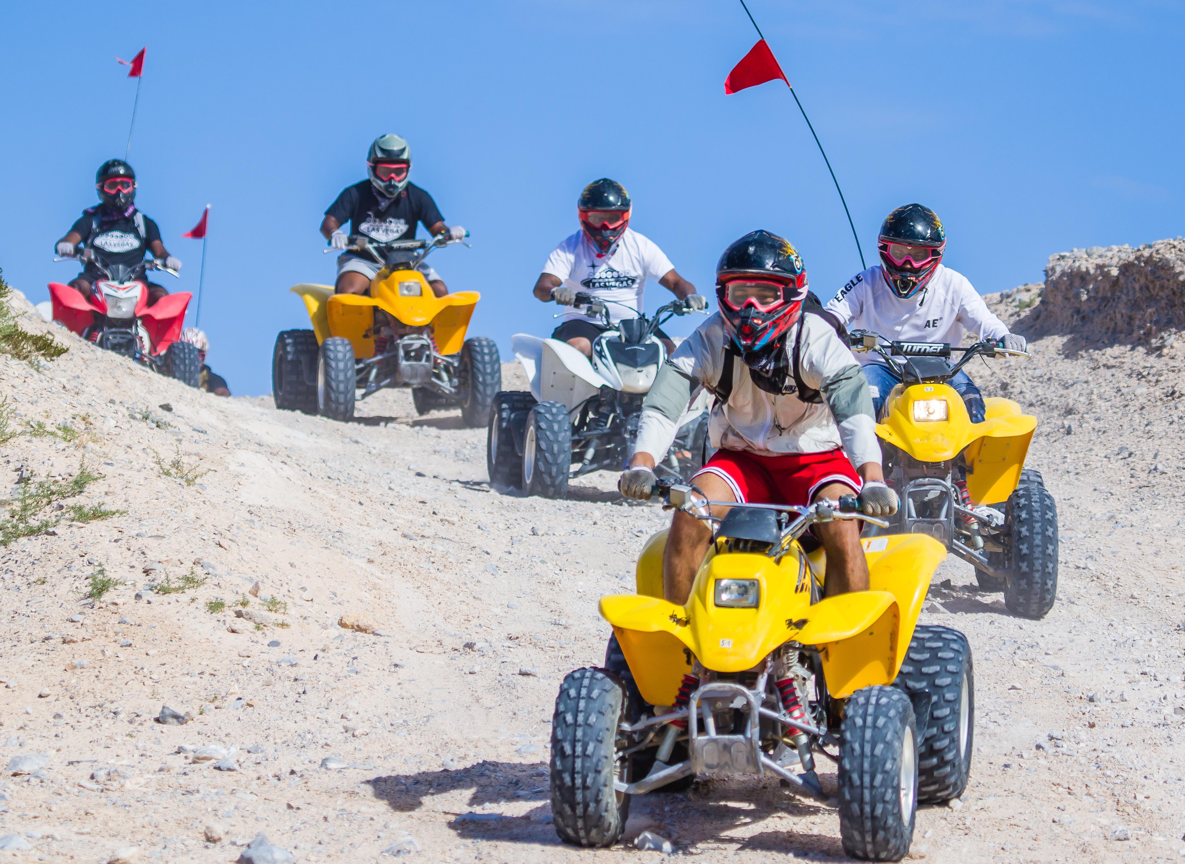 Five helmeted riders on yellow, red and white ATVs quad-biking down a sandy desert trail under a clear blue sky — off-road ATV tour in sand dunes.