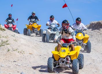Five helmeted riders on yellow, red and white ATVs quad-biking down a sandy desert trail under a clear blue sky — off-road ATV tour in sand dunes.