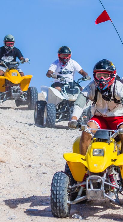 Five helmeted riders on yellow, red and white ATVs quad-biking down a sandy desert trail under a clear blue sky — off-road ATV tour in sand dunes.