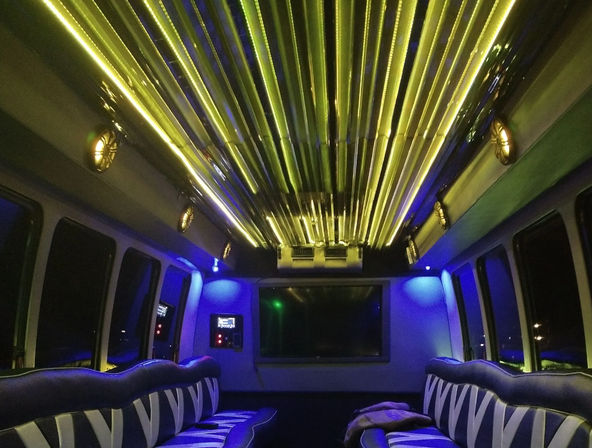 Luxury party bus interior with ribbed yellow LED ceiling, blue ambient lighting, striped leather bench seats and rear flat-screen TV at night