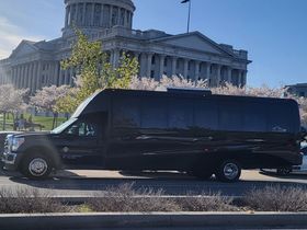 Sleek black passenger shuttle van parked on a city street with a domed neoclassical government building and blooming cherry trees in the sunny background.