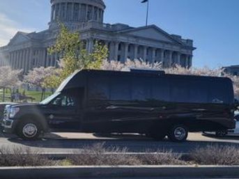Sleek black passenger shuttle van parked on a city street with a domed neoclassical government building and blooming cherry trees in the sunny background.
