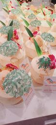 Rows of fresh young coconuts dressed as tropical drinks with green palm-print cocktail umbrellas, long leaves and pink orchids as beach-themed party favors on a white marble counter