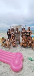 Group of women in swimsuits at a sandy beach bachelorette celebration by the ocean, posing with decorated coconut drinks and a macrame-style sign, pink inflatable float in the foreground.