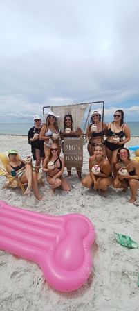 Group of women in swimsuits posing on a sandy beach by the ocean with a decorative bachelorette party sign, holding coconut drinks; pink inflatable float in the foreground and cloudy sky above.