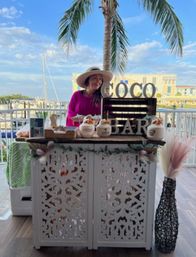 Person in a sun hat and pink top behind a white carved tropical coconut bar serving coconuts on a waterfront marina terrace with a palm tree and sailboats
