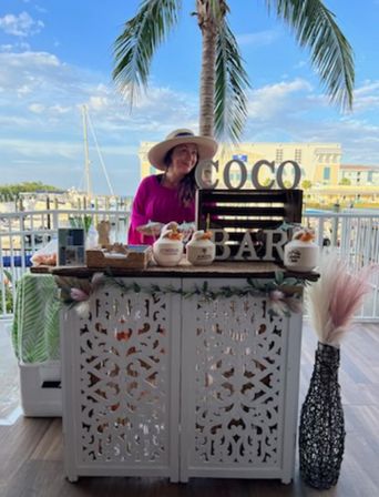 Person in a sun hat and pink top behind a white carved tropical coconut bar serving coconuts on a waterfront marina terrace with a palm tree and sailboats