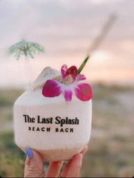 Hand holding a tropical coconut drink topped with a bright pink orchid and tiny umbrella, blurred beach sunset in the background.