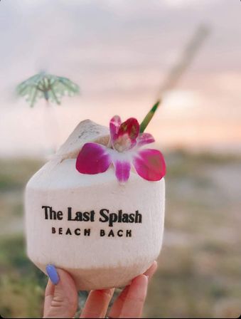 Hand holding a tropical coconut drink topped with a bright pink orchid and tiny umbrella, blurred beach sunset in the background.