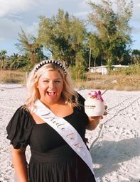 Smiling bride-to-be on a sunny sandy beach in a black dress and white sash, holding a coconut cocktail with a pink orchid and tropical trees in the background.