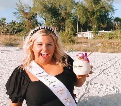 Smiling woman in a black dress wearing a 'Bride to Be' sash and beaded headband on a sunny sandy beach, holding a coconut cocktail garnished with a pink orchid with palm trees and dunes in the background