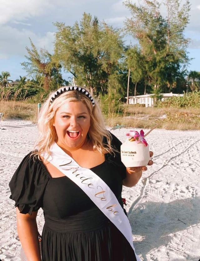 Smiling woman in a black dress wearing a 'Bride to Be' sash and beaded headband on a sunny sandy beach, holding a coconut cocktail garnished with a pink orchid with palm trees and dunes in the background