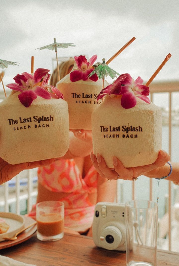 Three fresh coconuts garnished with pink orchids, paper umbrellas and straws raised in a beachside balcony toast — tropical drinks with a camera and coffee on the table.