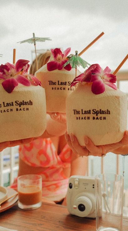 Three fresh coconuts garnished with pink orchids, paper umbrellas and straws raised in a beachside balcony toast — tropical drinks with a camera and coffee on the table.