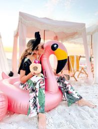 Person in floral beach pants and sunglasses kissing a large pink inflatable flamingo on white sand, holding a coconut tropical drink with orchid garnish beside pastel cabanas at sunset.