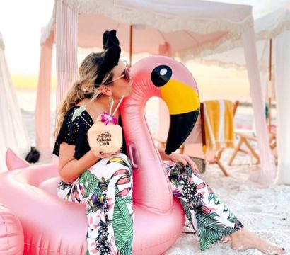 Person in floral beach pants and sunglasses kissing a large pink inflatable flamingo on white sand, holding a coconut tropical drink with orchid garnish beside pastel cabanas at sunset.